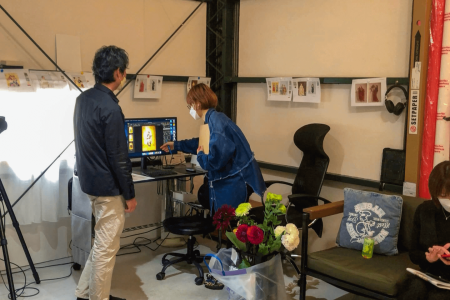 男性と女性が、写真撮影機材のある部屋でコンピューターのデスクで作業しています。女性はスクリーンを指差しており、近くにはカメラ付きの三脚が置かれています。手前には花と写真撮影用の背景があります。右側に座っているもう 1 人の人物は電話を使用しています。壁にはさまざまな写真がピンで留められています。