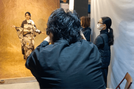 スタジオで、日本の伝統的な着物を着た女性がカメラマンにポーズをとっている。黒髪で眼鏡をかけたカメラマンは、カメラを彼女に向けている。2 人のアシスタントが近くに立っていて、1 人はマスクを着用し、観察している。背景は、質感のある金茶色のバックドロップである。